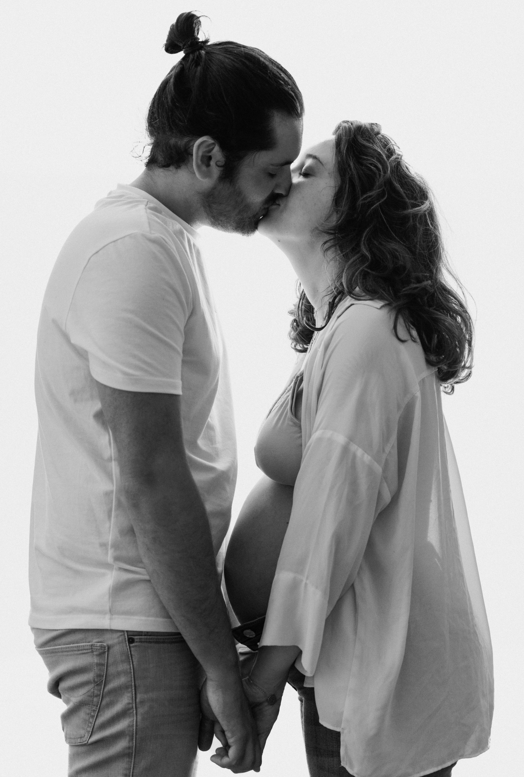 Tiphanie et Tristan s'embrassant, debout contre la fenêtre, Tristan la main sur le ventre de grossesse de Tiphanie, lors de leur séance photo de maternité pleine de tendresse à domicile à Bages (66).